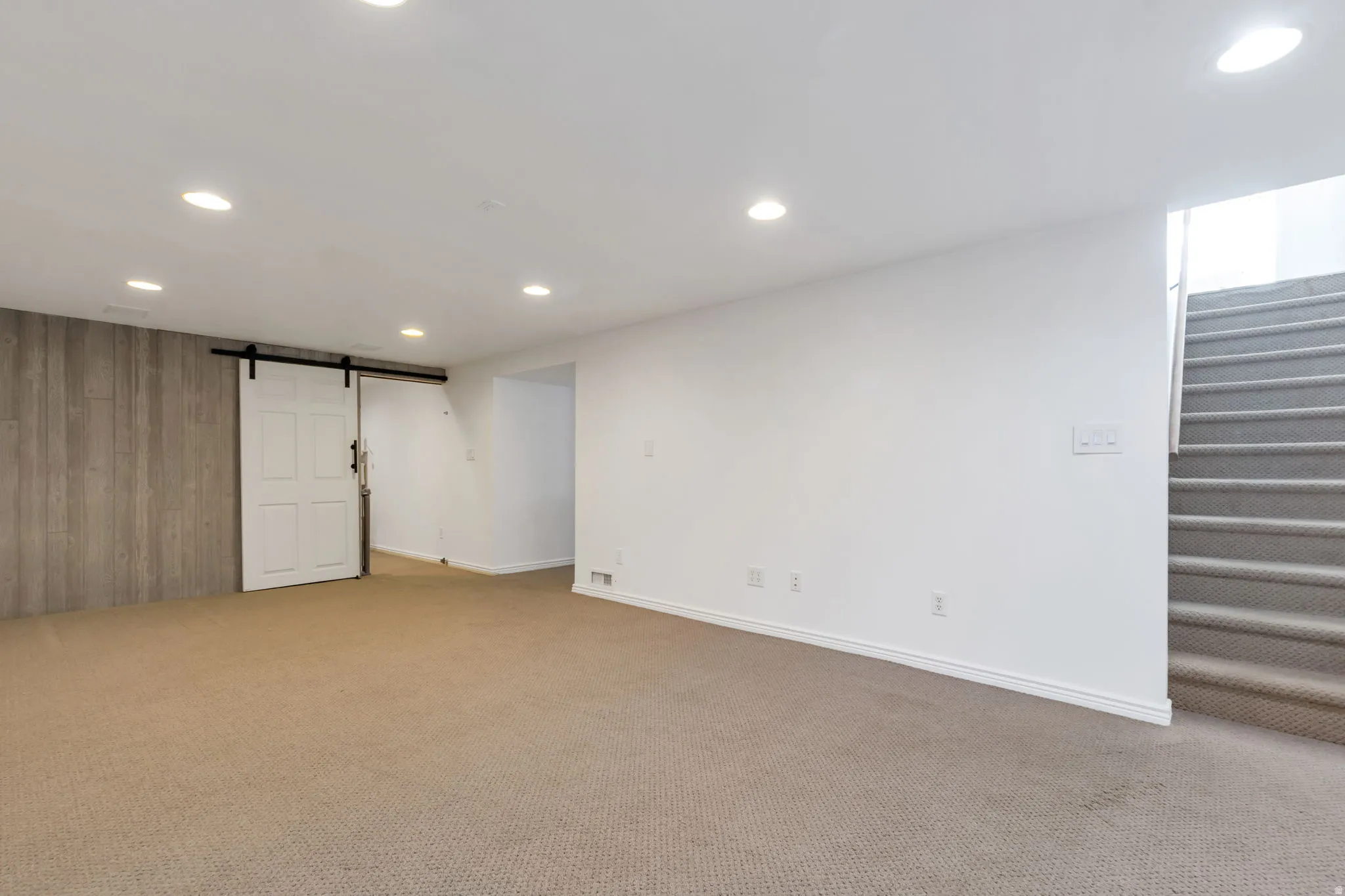Recessed lighting illuminates a spacious room featuring a neutral carpet, white walls, and a barn door with black hardware