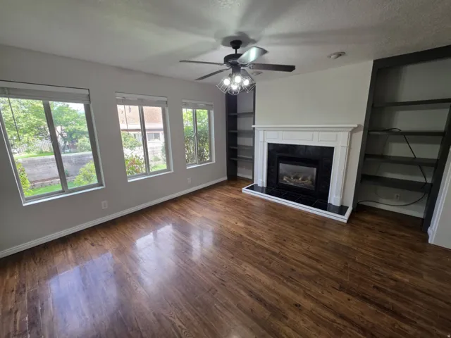 Living area featuring wood-finish flooring, a white mantel fireplace with black tile surround, a ceiling fan, and built-in shelving