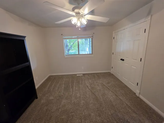 Neutral tone wall paint, textured carpet flooring, window with natural light, ceiling fan with integrated lighting, and white paneled doors