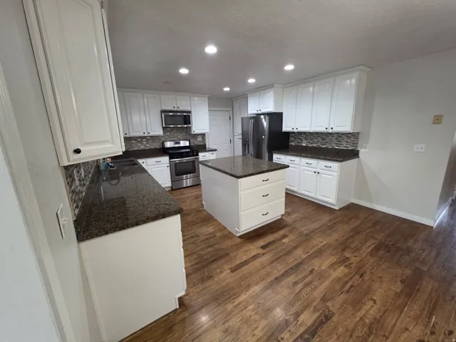 White shaker-style cabinetry, stainless steel appliances, dark stone countertops, mosaic tile backsplash, and a central island with storage