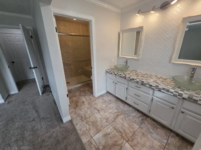 Dual vessel sinks set on a mosaic tile countertop, white cabinetry with dark hardware, a tiled shower with a built-in bench, tiled flooring, and decorative wall mirrors
