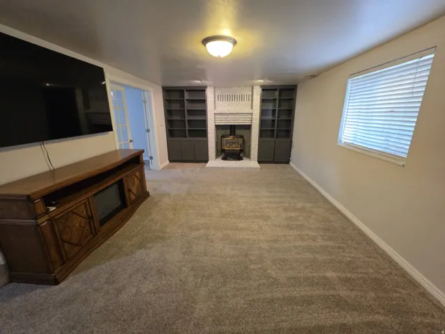 Plush carpeting, whitewashed brick fireplace with wood stove, built-in shelving, and a bright window