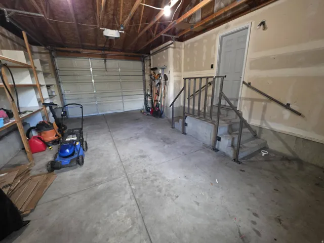 Concrete flooring, exposed wood beam ceiling, white overhead garage door, built-in wooden shelving, and interior door with metal railings