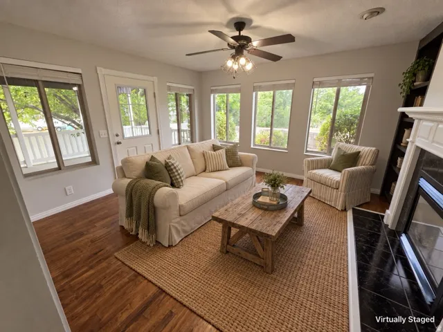 Living area featuring wood-finish flooring, multiple windows, a ceiling fan with integrated lighting, a built-in shelving unit, and a fireplace with a white mantel and dark tile surround