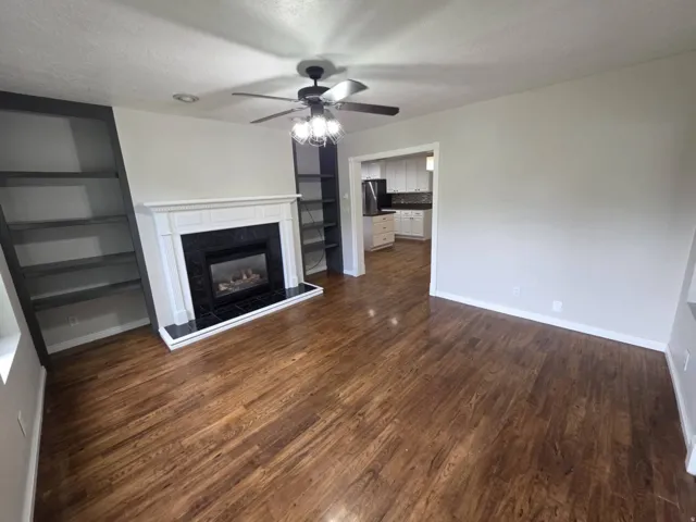 Living area featuring wood-finish flooring, a fireplace with a white mantel and black surround, built-in shelving, and a ceiling fan