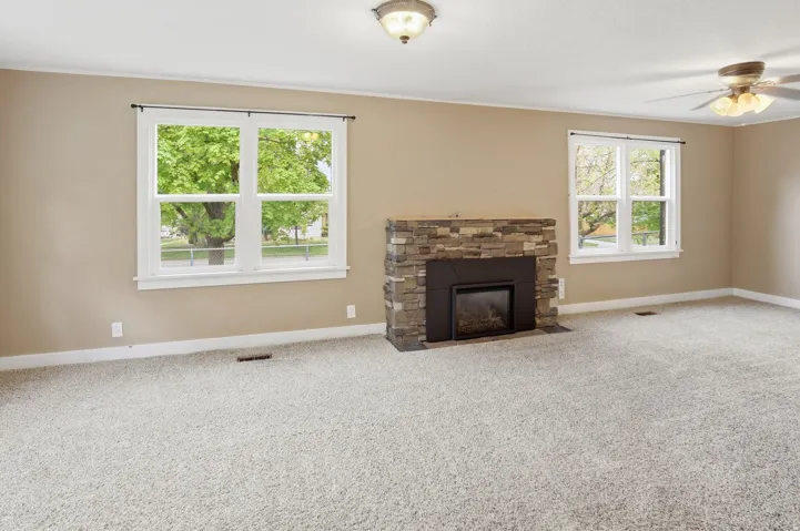 Carpeted room featuring a stone-clad fireplace, two double-hung windows, and a ceiling fan with integrated lighting