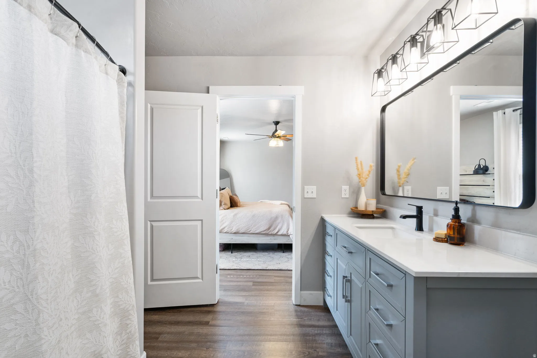 Wood-finish flooring, single vanity with light blue cabinetry, matte black hardware, rectangular mirror, and multi-light fixture