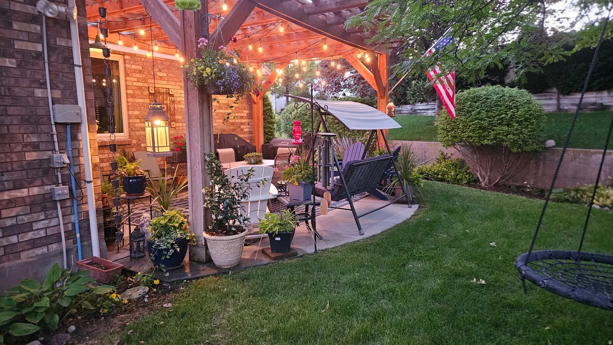 Covered patio with wood beam construction and ambient overhead lighting, overlooking a spacious lawn with mature landscaping and established shrubs