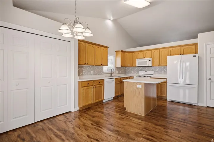 Vaulted ceiling kitchen with wood-finish flooring, light wood cabinetry, white appliances, and a central island