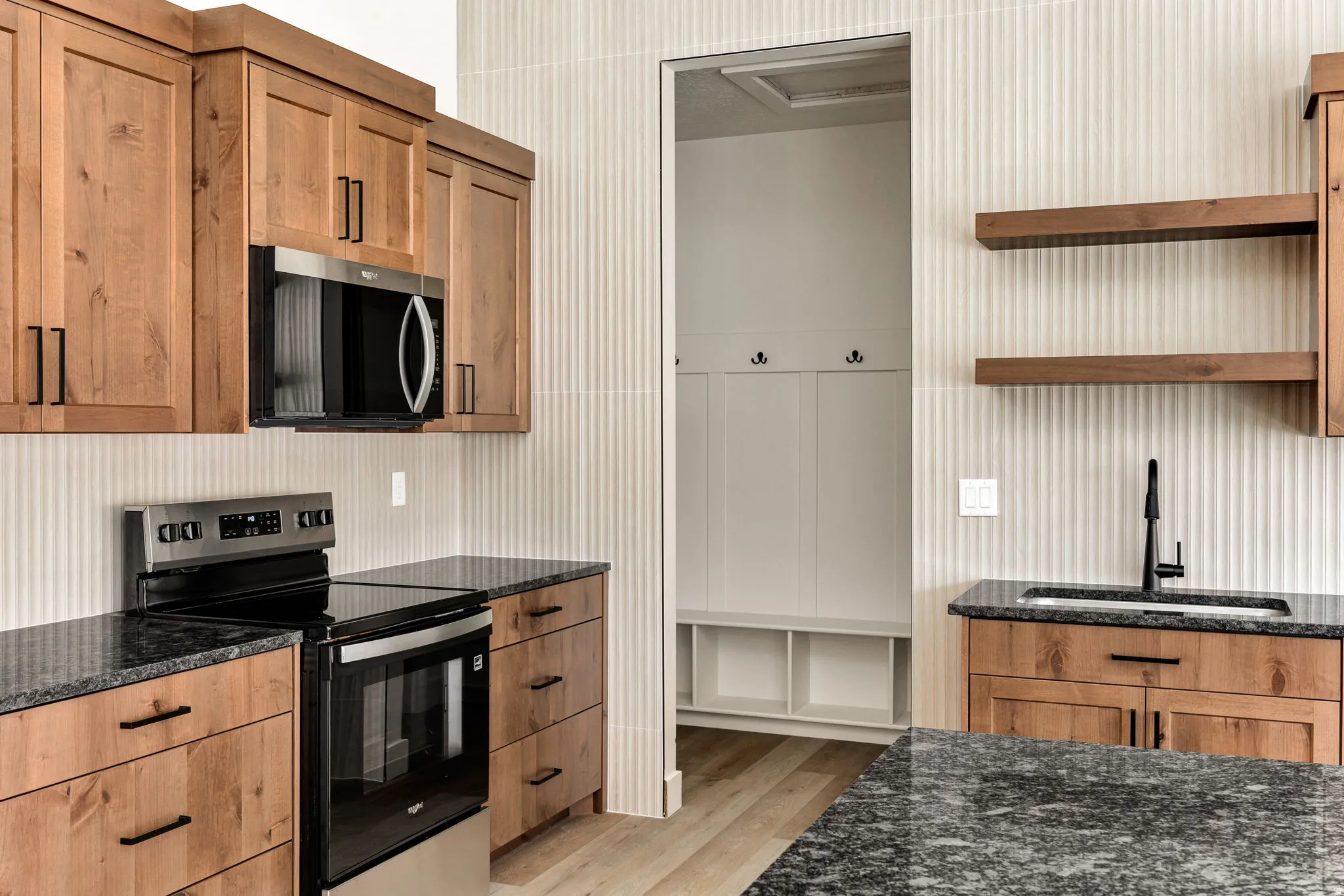 Wood-finish cabinetry and drawers, dark stone countertops, stainless steel appliances, a built-in boot bench, and wood-finish open shelving