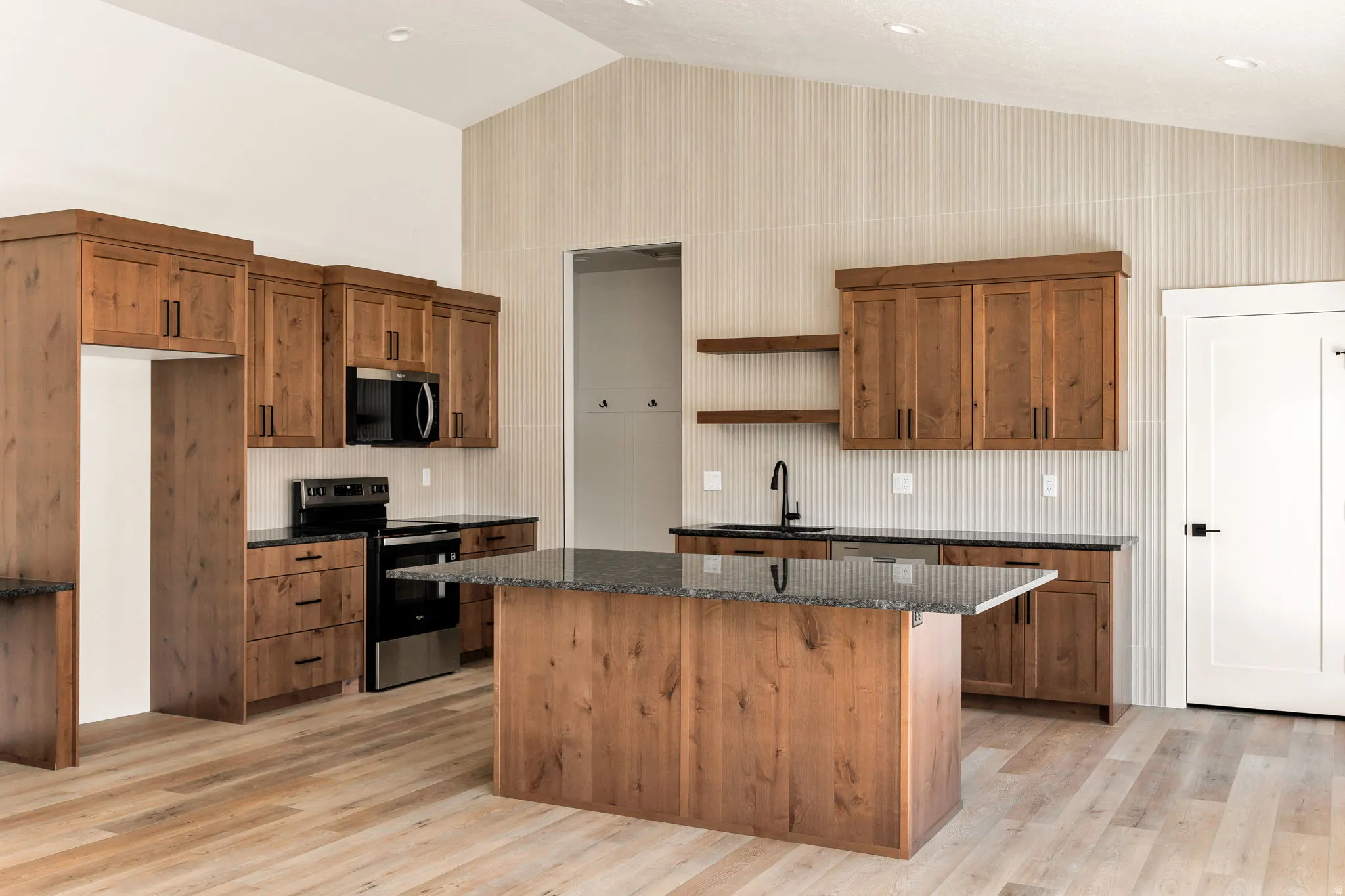 Vaulted ceiling kitchen featuring wood-finish cabinetry, a central island with a dark countertop, black appliances, and a black faucet