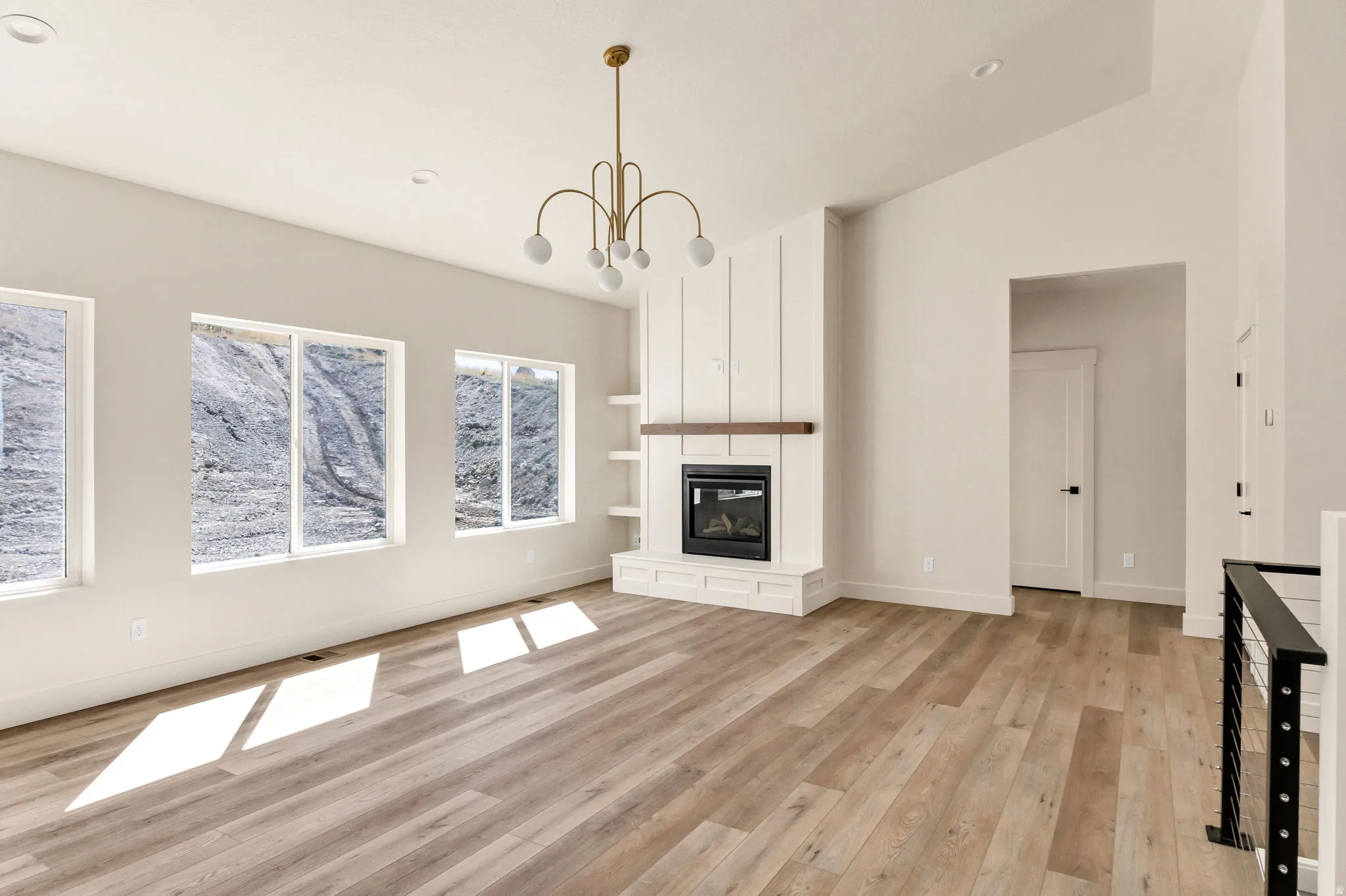 Vaulted ceiling with an attached brass chandelier, wood-finish flooring, a contemporary fireplace with a wood mantle, built-in shelving, and multiple windows