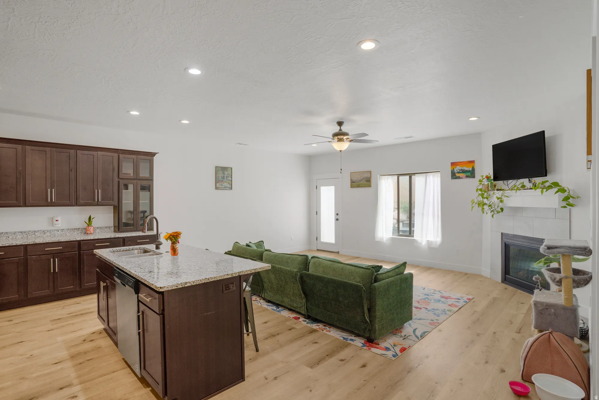 Open-concept living space with wood-finish flooring, a kitchen island with stone countertop, stainless steel appliances, and a fireplace with a mantel