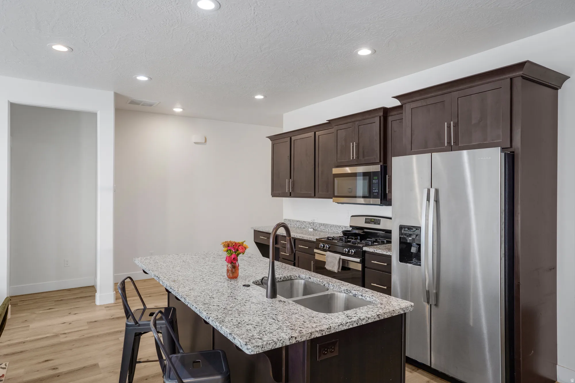 Kitchen featuring dark wood cabinetry, stainless steel appliances, a center island with a double basin sink, light-toned granite-style countertops, and recessed lighting