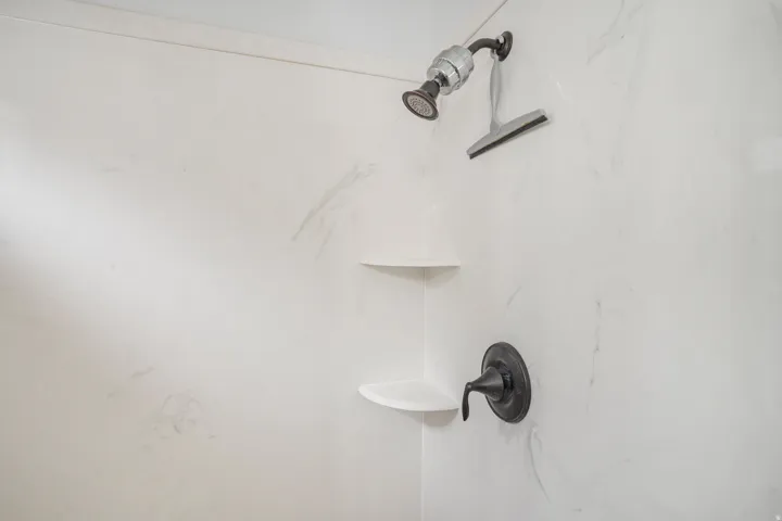 Shower featuring white stone-finish walls, a dark-finish shower head, and built-in corner shelving
