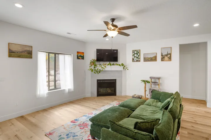 Recessed lighting, light wood-finish flooring, corner fireplace with tile surround, ceiling fan with integrated lighting, and window with white sheer curtains