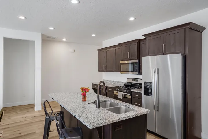 Kitchen featuring dark wood cabinetry, stainless steel appliances, a center island with a double basin sink, light-toned granite-style countertops, and recessed lighting
