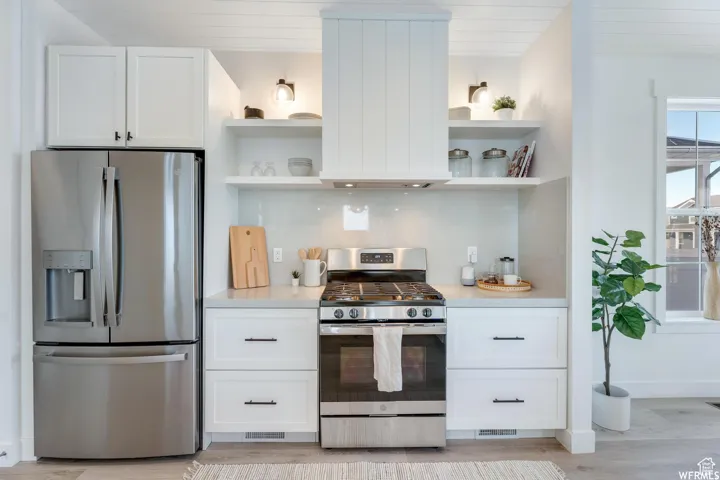 Stainless steel appliances, white shaker cabinetry, light-toned countertops, open shelving, and a built-in range hood