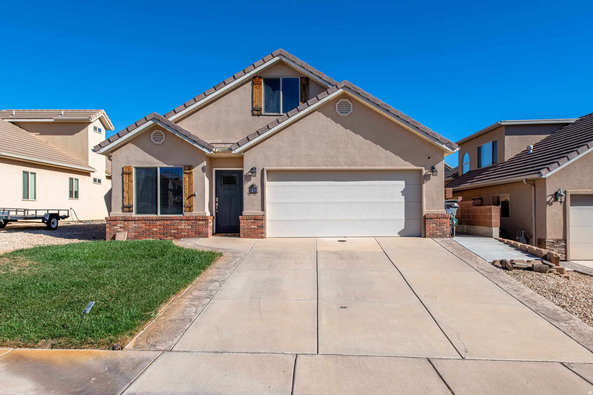 Stucco exterior with brick accents, wood-finish shutters, attached garage, concrete driveway, and front lawn