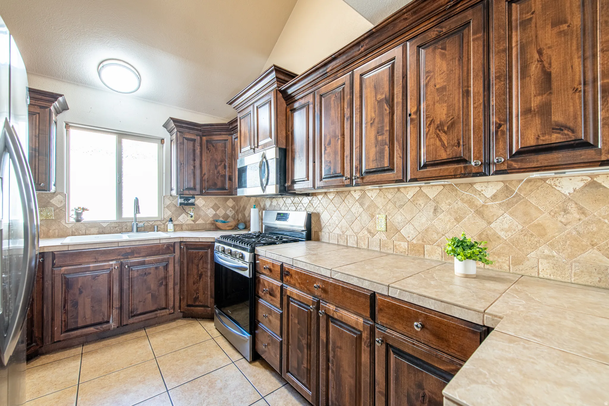 Kitchen featuring wood cabinetry, tile countertops, stainless steel appliances, a tile backsplash, and a vaulted ceiling