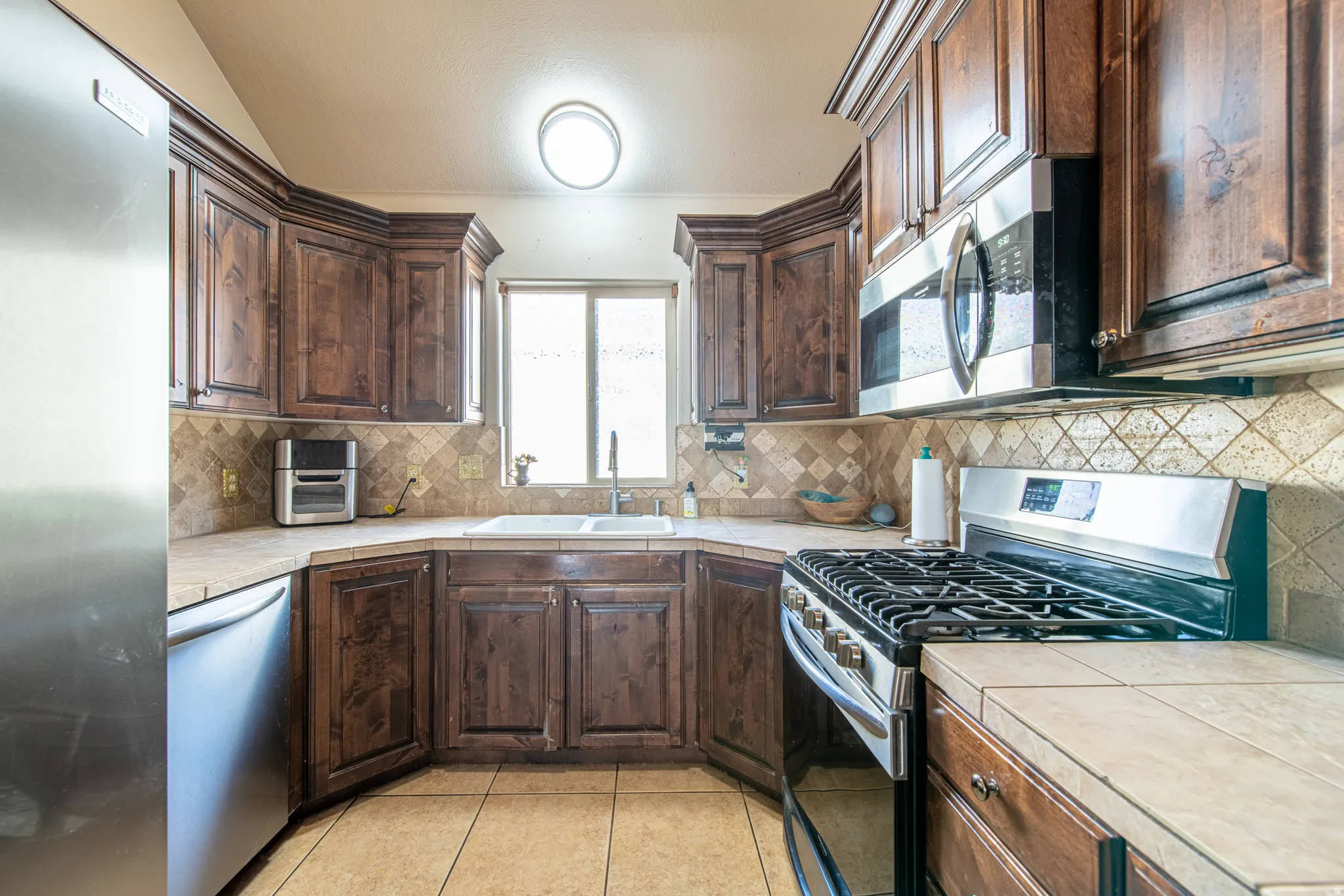 Kitchen featuring rich wood cabinetry, tile countertops, a coordinating tile backsplash, and stainless steel appliances including a range and microwave