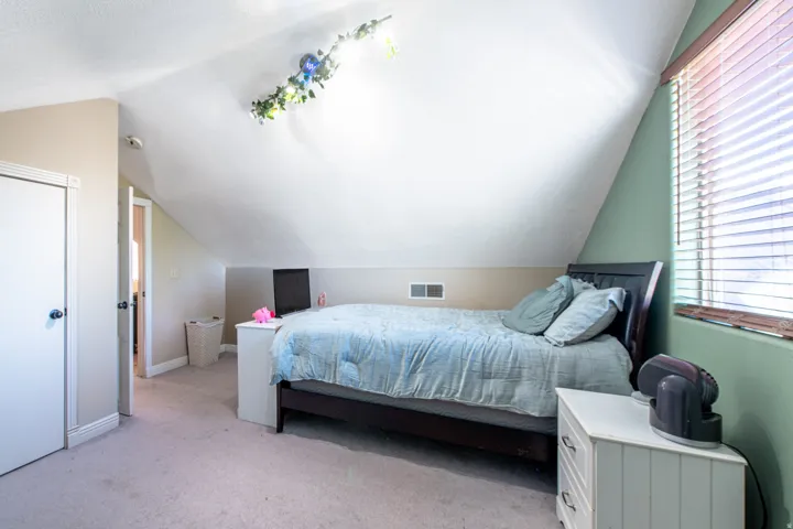 Angled ceiling, window with blinds, light-colored wall-to-wall carpeting, white interior door, and a wall vent