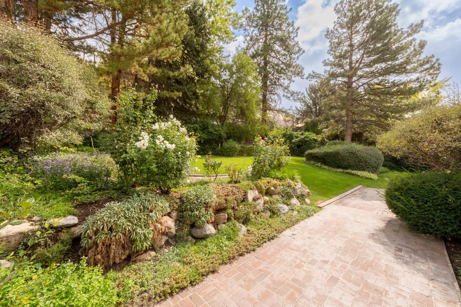 Expansive outdoor space featuring a brick-finish walkway, terraced landscaping with rock retaining walls, mature trees, and manicured lawns