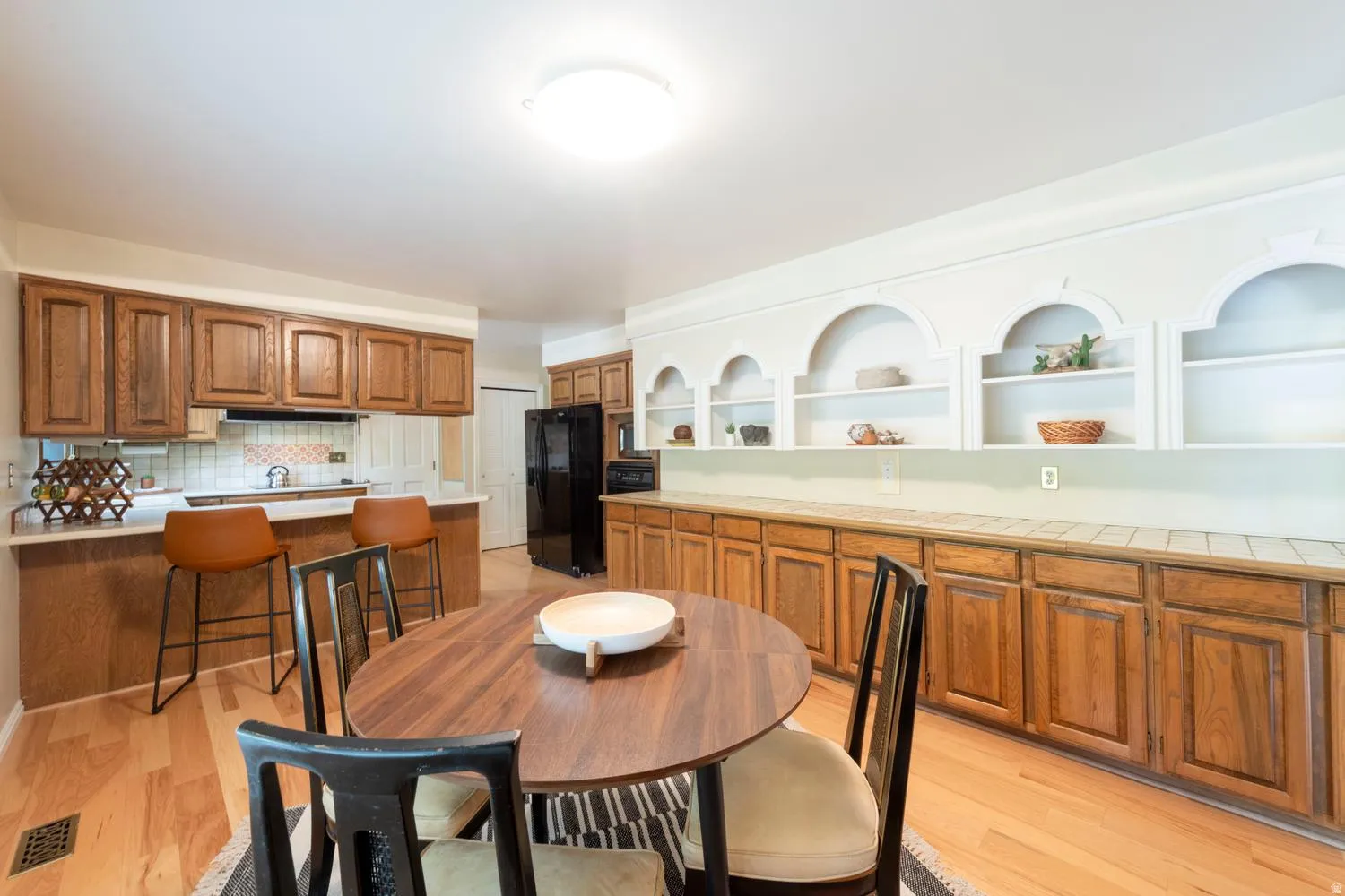 Wood-finish flooring, wood cabinetry, built-in shelving with arched accents, tiled countertop, and a kitchen island with seating