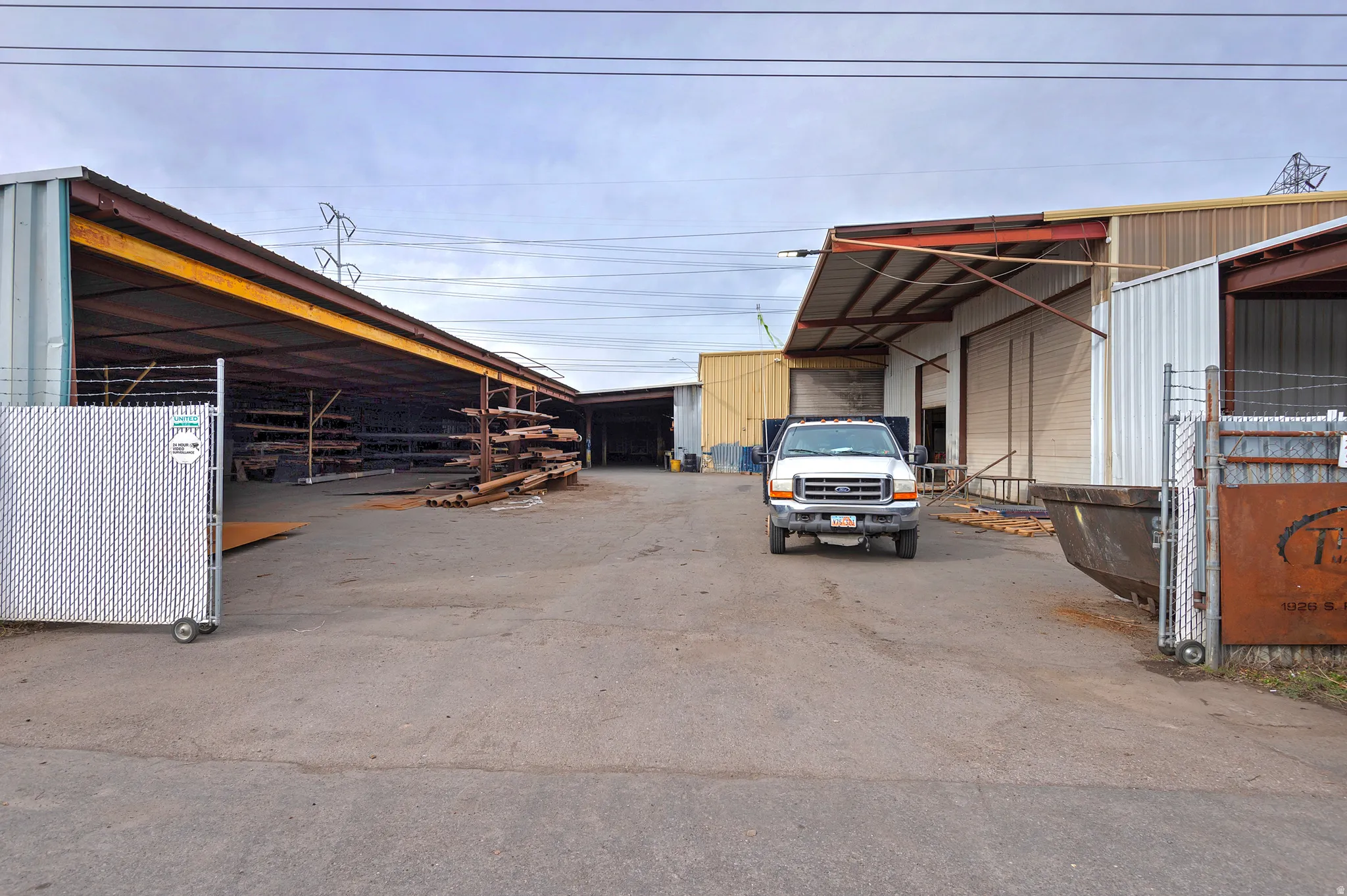 Paved lot with multiple warehouse structures featuring roll-up doors, an open-sided storage area with metal shelving, and perimeter chain link fencing with a gate