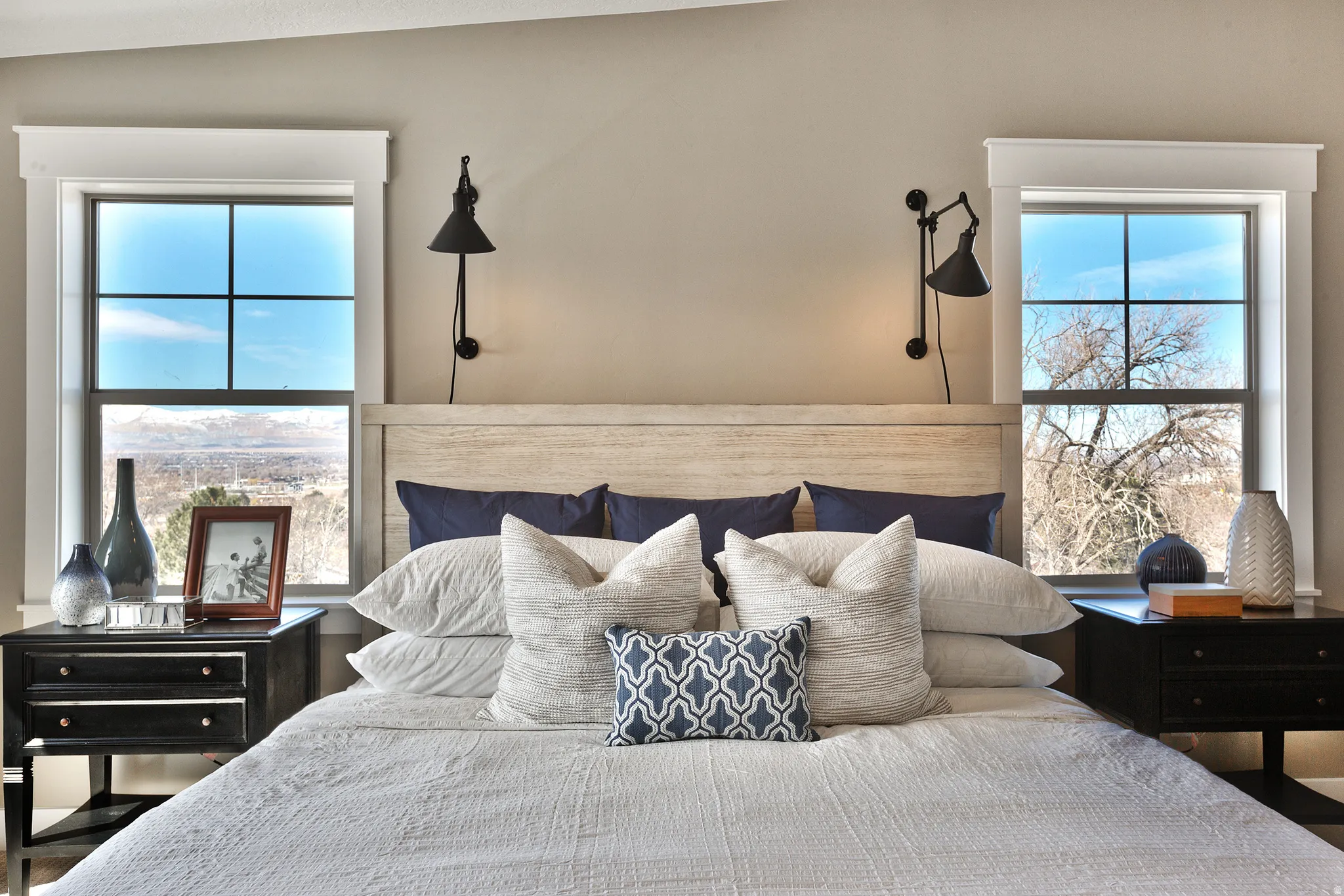 Primary Bedroom, Dual windows with white trim, a light wood-finish headboard, and wall-mounted sconces