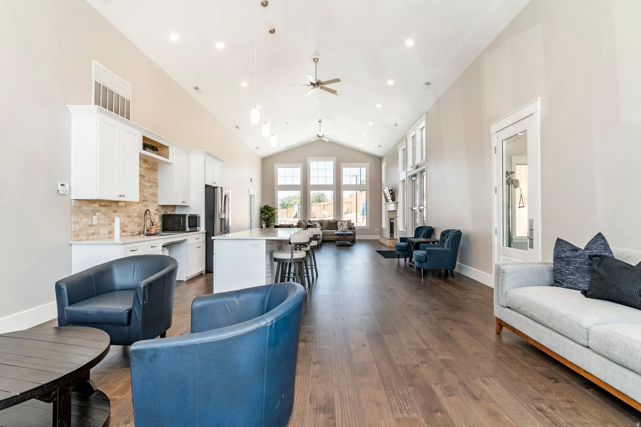 Vaulted ceilings, wood-finish flooring, a kitchen area with white cabinetry and stone-look backsplash, an island with seating, and tall windows