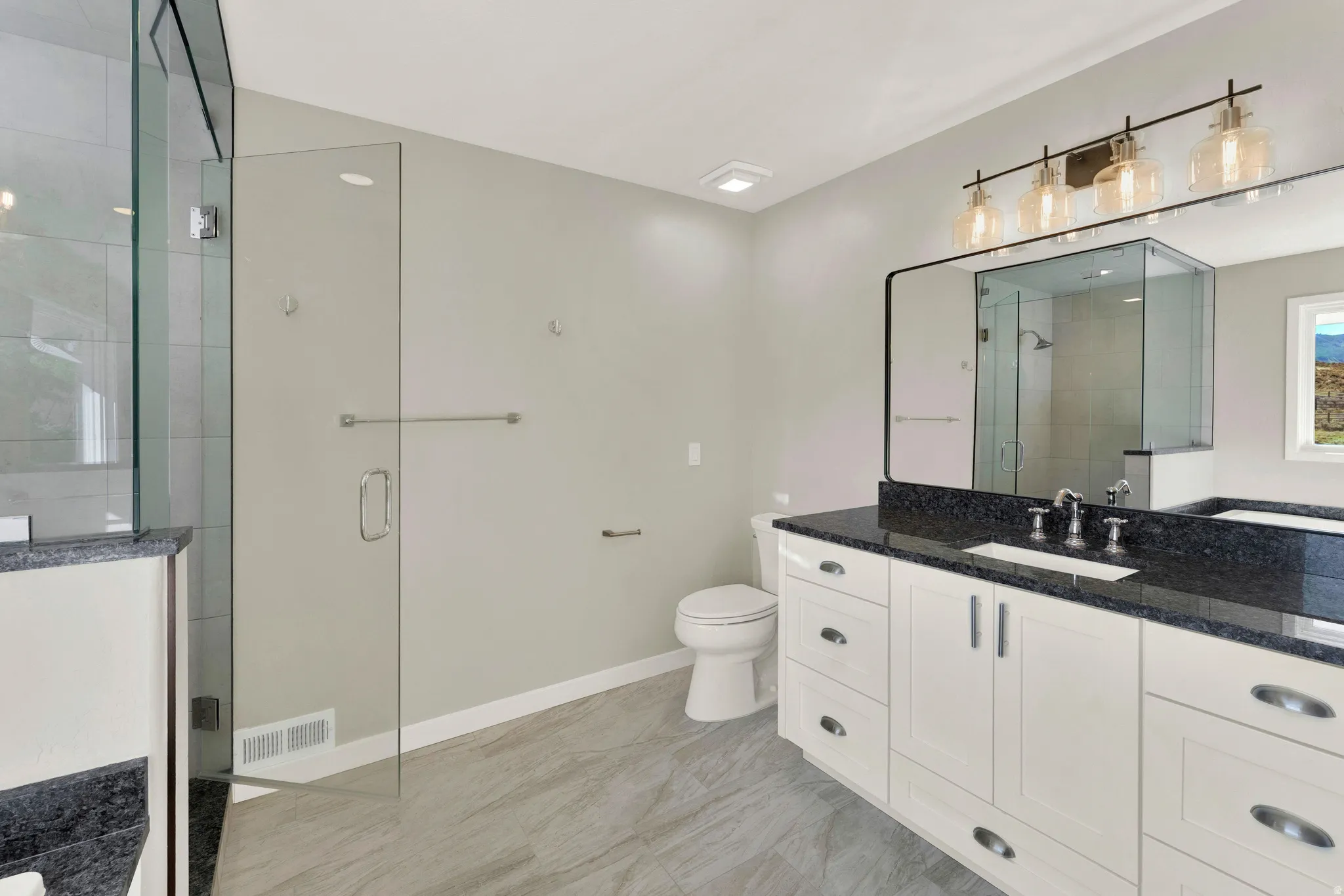 Frameless glass shower enclosure, dual vanity with dark countertop, white vanity cabinetry, wall-mounted light fixture, and wood-finish flooring