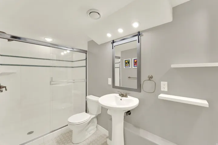 Shower with sliding glass doors, pedestal sink, wall-mounted mirror with integrated lighting, and two floating wall shelves