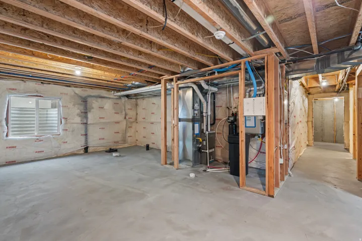 Unfinished basement with exposed wood joists, concrete flooring, window with horizontal blinds, framed utility area, and insulation on wall surfaces