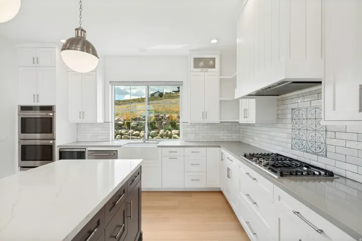 White shaker cabinetry, light wood-finish flooring, built-in double ovens, farmhouse sink, and a decorative tile backsplash