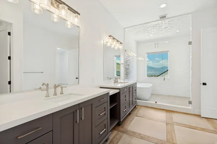 Dual vanity with white countertops and dark wood cabinetry, freestanding bathtub, glass-enclosed shower with white subway tile, and light-toned floor tiling