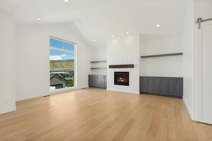 Vaulted ceiling with recessed lighting, large picture window, light wood-finish flooring, built-in fireplace with wood mantel, and integrated cabinetry with shelving