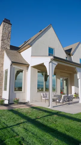 Stone chimney, light-colored stucco exterior, covered patio, multiple windows, and a manicured lawn