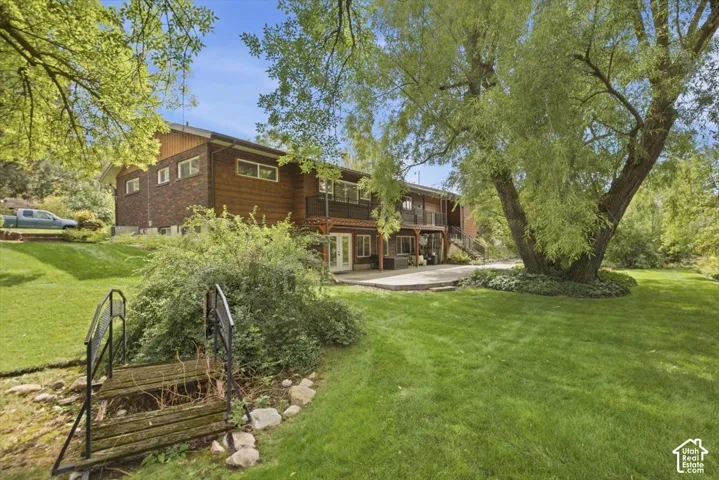 Expansive rear yard featuring mature trees, a manicured lawn, a large paved patio, and a wood and brick exterior