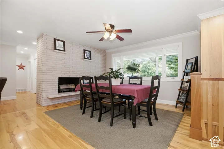 Wood-finish flooring, white brick fireplace with hearth, white window trim, natural wood paneling, and a ceiling fan with integrated lighting