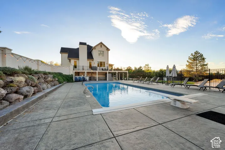 In-ground swimming pool with diving board, stamped concrete patio, tiered landscaping with stone retaining wall, and a raised deck with metal railing