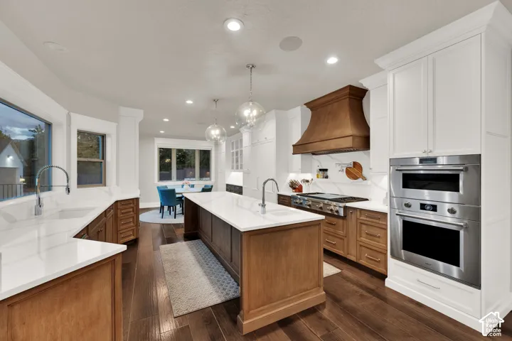 Kitchen featuring a large central island, white countertops, built-in stainless steel appliances, a wood-finish range hood, and wood-finish flooring