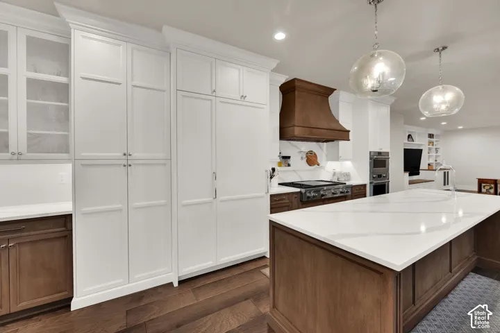 Expansive kitchen island with a light-toned countertop, wood-finish flooring, white cabinetry, integrated appliances, and pendant lighting