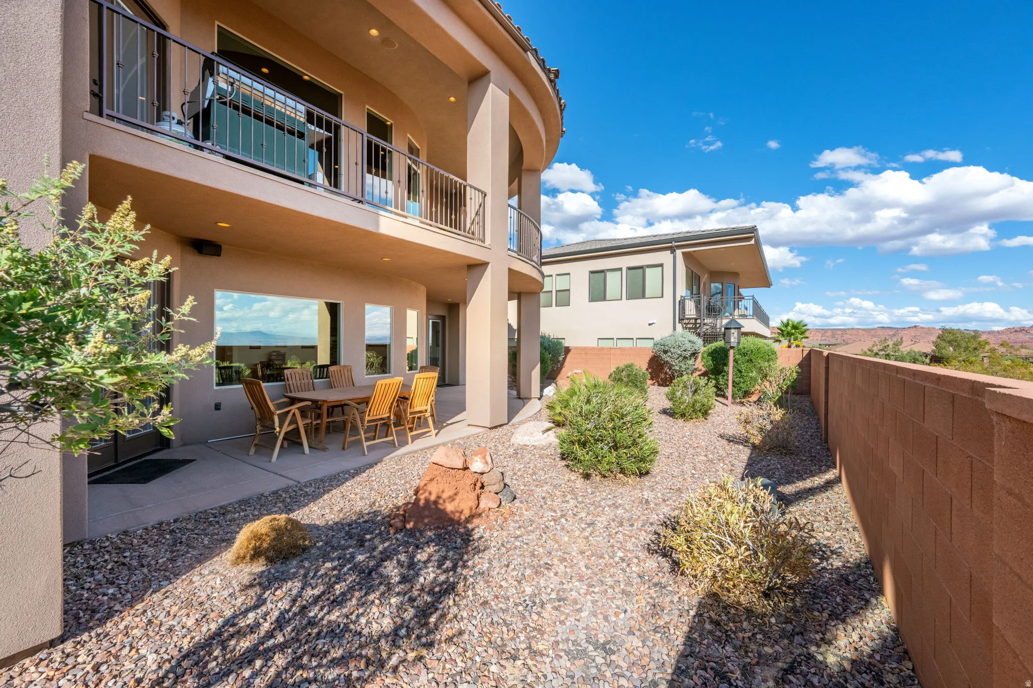 Curved exterior architecture, ground-level patio, upper-level balcony with metal railing, expansive windows, and a landscaped rock garden