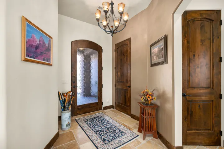 Arched wood-finish entry door with textured glass, complementing solid wood-finish interior doors, stone tile flooring, and a wrought iron chandelier