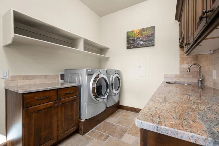 Laundry area with stone tile flooring, granite countertops, wood cabinetry, and a stainless steel sink with a gooseneck faucet