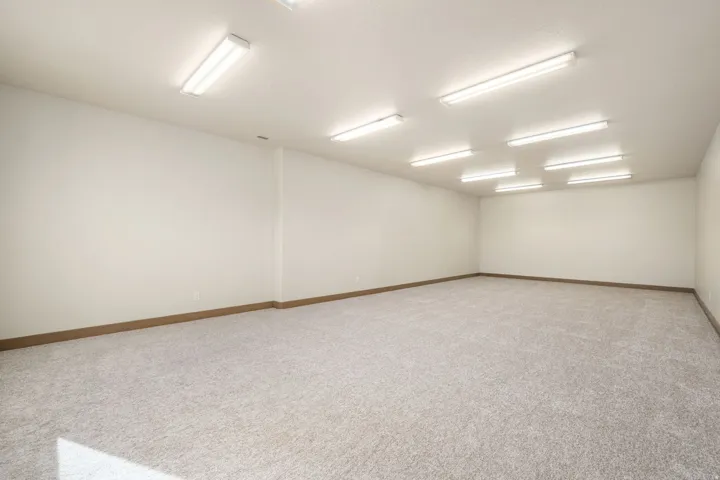 Spacious room featuring neutral carpet flooring, light-colored walls, wood-finish trim, and ample overhead fluorescent lighting