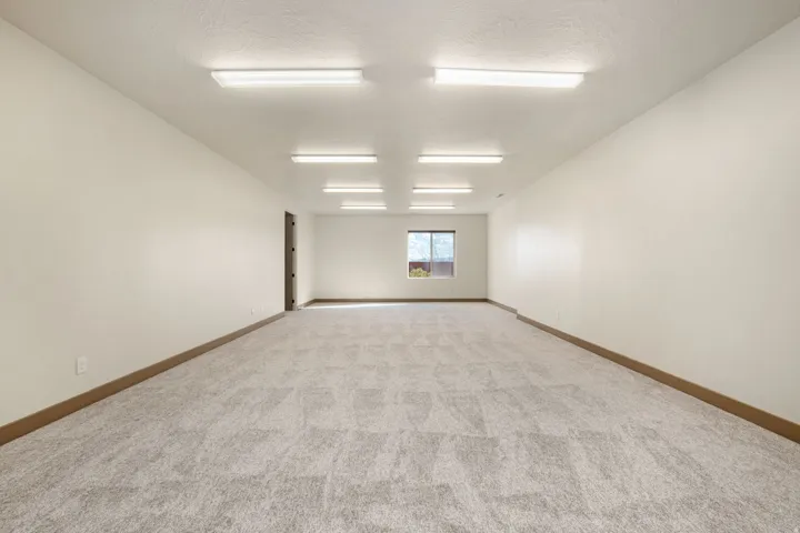 Expansive interior space featuring light-toned carpeting, ample overhead lighting, a single window, and a dark-toned door frame