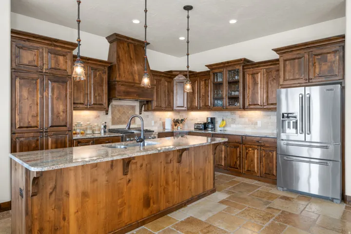Wood cabinetry throughout, stone tile flooring, granite countertops, stainless steel refrigerator, and pendant lighting
