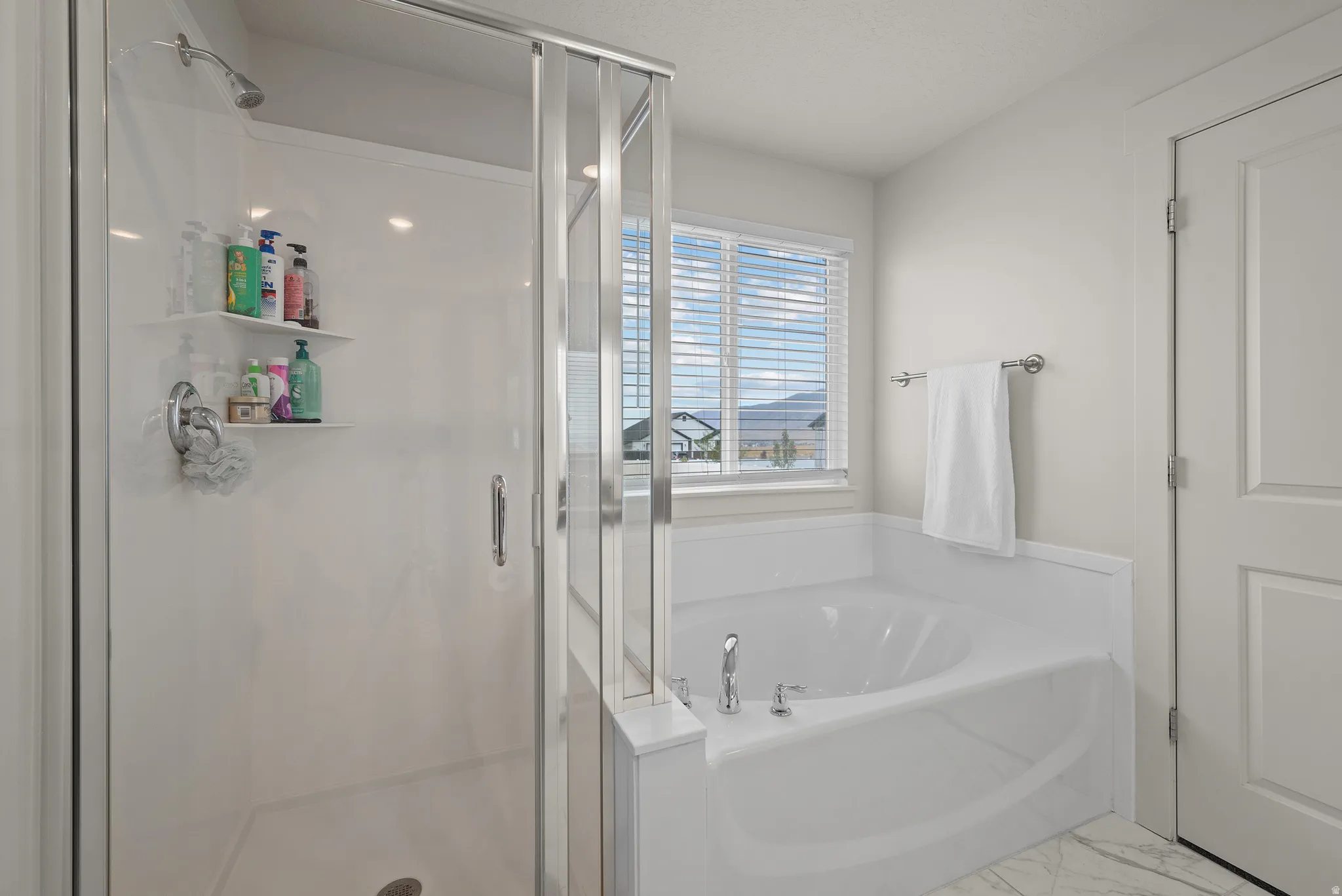 Walk-in shower with clear glass enclosure and built-in shelving, white drop-in bathtub with deck-mounted fixtures, and a window with blinds